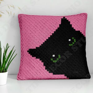 May include: A square crochet throw pillow featuring a vibrant pink background and a large, black, pixelated cat silhouette. The cat design includes two distinct green eyes with white pupils, rendered in a textured, hand-stitched yarn pattern.