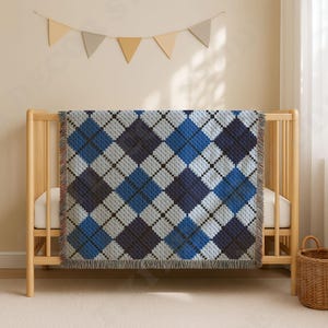 Op de afbeelding: Een geweven plaid met een herhalend argyle-patroon in tinten marineblauw, middenblauw en lichtgrijs. De deken heeft een textuurafwerking met zichtbare franjes langs de verticale randen en hangt over een ledikant van lichtgekleurd hout.