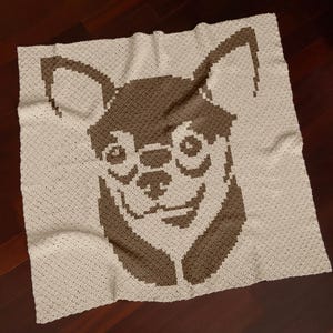 Può includere: Una coperta quadrata all'uncinetto color crema con un disegno di un muso di cane chihuahua marrone. Il muso del cane è posizionato centralmente, con occhi, naso e bocca sorridente dettagliati. La coperta è su una superficie di legno scuro.