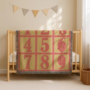 Könnte beinhalten: Eine gelbe Babydecke mit einem Rastermuster, das die Zahlen 4, 5, 6, 7, 8 und 9 in Koralle zeigt. Die Decke hat einen Fransenrand und liegt über einem Holzbettchen.