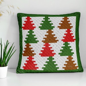 May include: A square, crocheted pillow with a green border. The pillow features a repeating pattern of Christmas trees in red, green, and brown on a white background. The pillow is a decorative item.