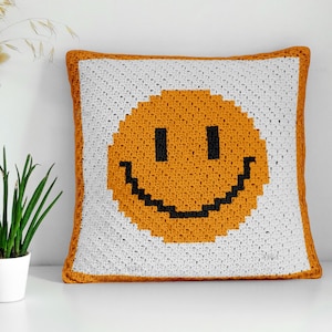 Può includere: Cuscino quadrato all'uncinetto con una grande faccina sorridente pixelata. Il viso è arancione con occhi neri e un sorriso nero, su sfondo bianco. Il cuscino ha un bordo arancione.