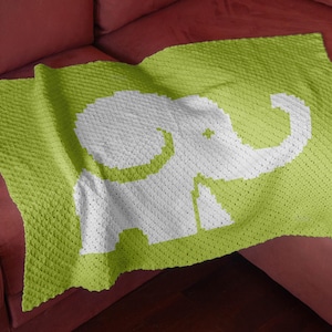 May include: A lime green crocheted blanket with a white elephant design. The elephant is centered on the blanket and has large ears and a curled trunk. The blanket is folded on a red sofa.
