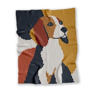 May include: A crocheted blanket with a beagle dog design. The dog's head and upper body are shown in white, brown, black, and gold. The blanket has a textured look and is folded, displaying the dog's profile against a colour-blocked background.