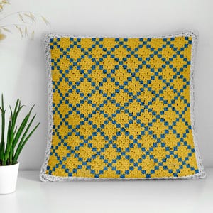 May include: Square crochet cushion featuring a yellow and blue geometric pattern. The cushion has a white border. The cushion is placed on a white surface. Decorative item for a sofa or bed.