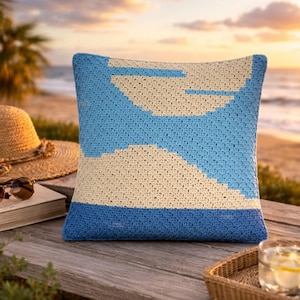 May include: Square crochet pillow with a beach-themed design. The pillow features a blue and cream color scheme, with a geometric pattern resembling waves and clouds. The pillow is placed on a wooden surface near the beach.