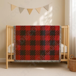 May include: A rectangular woven throw blanket featuring a bold red and black buffalo plaid pattern with a textured, diagonal weave and fringed edges, draped over the wooden slats of a light-colored crib in a nursery setting.
