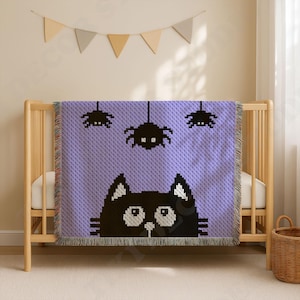 Puede incluir: Una manta tejida con fondo morado claro que presenta una cara de gato negro en la parte inferior y tres arañas negras colgando del borde superior. La manta tiene un borde con flecos y un diseño pixelado en toda su superficie.
