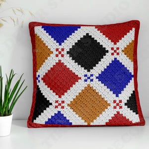 May include: A square crochet pillow cover featuring a geometric argyle pattern. The design consists of diamond shapes in red, blue, black, and tan, separated by white crocheted lines. The entire pillow is bordered by a solid red crocheted edge.