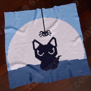 Puede incluir: Una manta de ganchillo cuadrada con un diseño de pixel art de un gato negro sentado frente a una gran luna llena blanca sobre un cielo azul claro. Una pequeña araña negra cuelga de un hilo sobre la cabeza del gato, sobre una base azul oscuro.