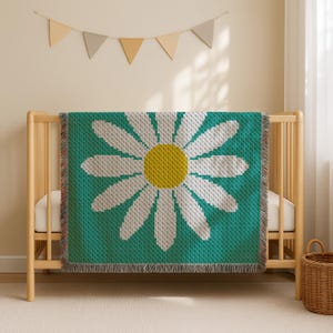 May include: A teal baby blanket with a large daisy design. The daisy has white petals and a yellow center. The blanket is draped over a wooden crib. The crib is in a room with a beige wall and a white curtain.