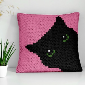 May include: A square, pink crochet pillow featuring a black cat design. The cat's face is a geometric shape with green eyes. The pillow is a decorative item, likely for a bed or couch.