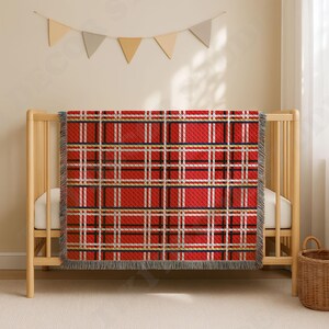 May include: A rectangular woven throw blanket featuring a red, white, black, and gold plaid pattern with a fringed edge. The blanket is draped over a light-coloured wooden crib, displaying a classic tartan design with intersecting horizontal and vertical lines.