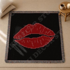 Puede incluir: Una manta negra con una imagen grande y pixelada de labios rojos. La manta tiene un borde con flecos. Los labios son un diseño llamativo y audaz. La manta se muestra sobre una superficie de color neutro.