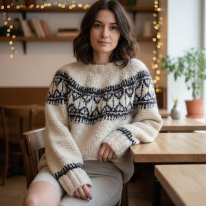 Può includere: Una donna indossa un maglione color crema con un motivo geometrico nero sul petto e sulle maniche. Il maglione ha una maglia spessa e strutturata. È seduta a un tavolo di legno in un bar.