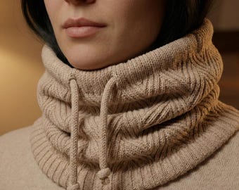 May include: A beige knit cowl neck warmer with a drawstring closure. The neck warmer is made of a soft, textured material and is designed to be worn around the neck for warmth and style. The cowl neck is a neutral color.