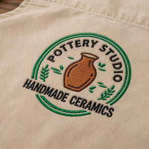 May include: A beige fabric apron featuring an embroidered circular logo. The design includes a brown ceramic vase in the center, surrounded by green leaf accents and the black text POTTERY STUDIO HANDMADE CERAMICS arranged in a circular pattern.