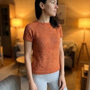 May include: A short-sleeved, orange knit top with a crew neck. The top is made of a textured yarn and has a ribbed hem and cuffs. The model is wearing gray shorts. The background is a cozy living room.