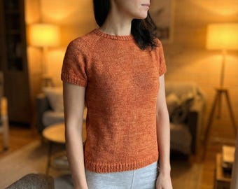 Pode incluir: Uma blusa de malha laranja com mangas curtas e gola redonda. A blusa é feita de um fio texturizado e tem bainha e punhos canelados. A modelo está usando shorts cinza. O fundo é uma sala de estar aconchegante.