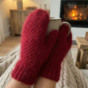 Puede incluir: Primer plano de un par de mitones rojos tejidos, con puño acanalado, sosteniendo una taza beige. Los mitones están hechos de un hilo suave y texturizado. Un fondo con una chimenea y una manta.