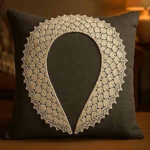 May include: A cream-coloured lace collar with a geometric design rests on a dark grey cushion. The collar is shaped like a teardrop, with the lace forming a decorative border. The cushion provides a contrasting backdrop, emphasising the intricate details of the lace.