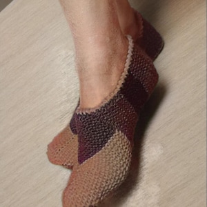Pode incluir: Um par de pantufas de malha em tons de bege, roxo e bordô. As pantufas têm um padrão de malha texturizado e uma borda contrastante ao redor da abertura. As pantufas são projetadas para cobrir o pé.