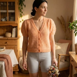 May include: A peach-colored, short-sleeved cardigan with a V-neck and button closure. The cardigan has a textured knit design and is paired with gray shorts. The person is holding a small bouquet of dried flowers.