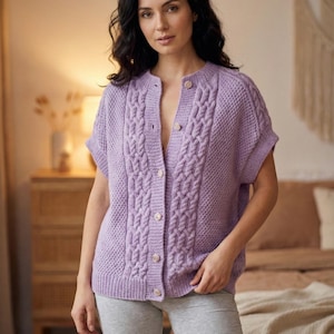 Puede incluir: Un cárdigan lila de manga corta con un diseño de punto trenzado en la parte delantera y en las mangas. El cárdigan tiene cuello redondo y se abrocha con botones. Los botones son de color beige claro. La persona lleva unos pantalones cortos grises.
