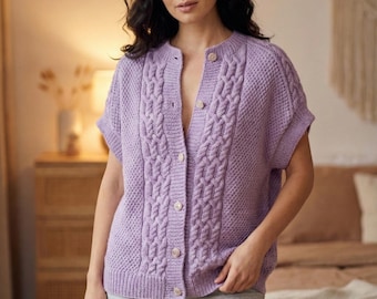 Pode incluir: Um cardigan lilás de manga curta com um padrão de malha entrançada na frente e nas mangas. O cardigan tem decote redondo e é abotoado. Os botões são de cor bege claro. A pessoa está a usar calções cinzentos.