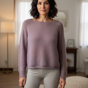 May include: A light purple sweater with a boat neck and long sleeves. The sweater is paired with gray biker shorts. The woman in the image has dark hair and is standing in a well-lit room.