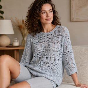 Può includere: Un maglione azzurro chiaro a maglia aperta con scollo tondo e maniche a tre quarti. Il maglione presenta un motivo ovale ripetuto. La persona indossa anche pantaloncini grigi. Il maglione è una maglia leggera e ariosa, perfetta per la stratificazione.