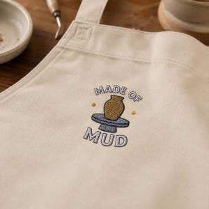 May include: A beige canvas apron featuring an embroidered design of a brown pottery vase on a blue potter's wheel. The text "MADE OF MUD" is stitched in grey thread above and below the central graphic, accented by two small yellow dots.