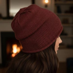 Könnte beinhalten: Eine bordeauxfarbene Strickmütze. Die Mütze ist aus dickem, strukturiertem Garn gefertigt und hat einen umgeschlagenen Rand. Die Mütze hat eine tiefrote Farbe und wirkt warm und gemütlich. Die Mütze wird von einer Person getragen.