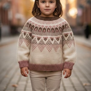 May include: A child's ivory sweater with a geometric pattern in shades of brown and pink. The sweater has a round neckline and ribbed cuffs and hem. The child is also wearing beige pants.