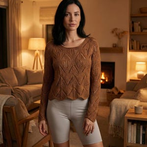 May include: A woman wearing a brown, long-sleeved sweater with a unique open-knit pattern. The sweater has a rounded neckline and a scalloped hem. She is also wearing gray biker shorts. The background shows a cozy living room setting.