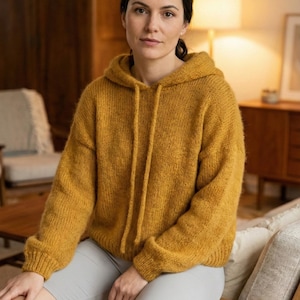 Puede incluir: Suéter con capucha amarillo mostaza con cordones. El suéter está hecho de una tela de punto suave y texturizada. La persona de la imagen lleva pantalones cortos grises. El fondo incluye una mesa de madera, una silla y una lámpara.