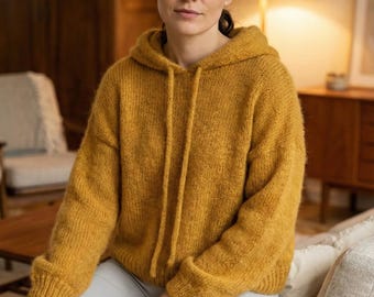 May include: Mustard yellow hooded sweater with drawstrings. The sweater is made of a soft, textured knit fabric. The person in the image is wearing gray shorts. The background includes a wooden table, chair, and a lamp.