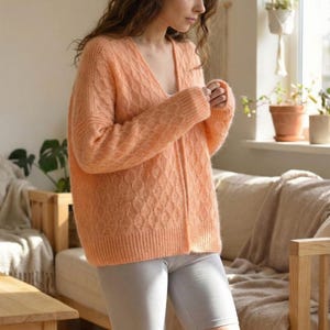May include: A peach-colored, long-sleeved cardigan with a V-neck and a textured knit pattern. The cardigan is paired with gray biker shorts. The outfit is casual and comfortable, suitable for everyday wear.