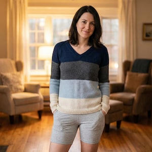 Può includere: Una persona indossa un maglione con scollo a V con righe orizzontali blu navy, grigio, azzurro e crema. Indossa anche pantaloncini grigi. Il maglione ha una vestibilità ampia. Lo sfondo comprende due poltrone e una finestra.