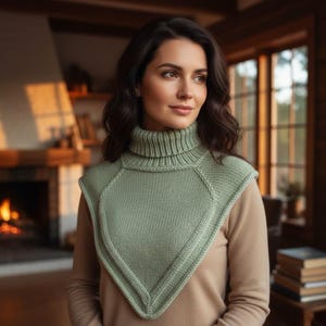 May include: A light green knitted cowl with a turtleneck design. The cowl features a diamond-shaped front panel and is worn over a tan-colored sweater. The image is set in a warm, cozy interior with a fireplace in the background.