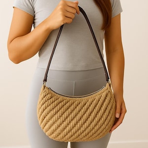 May include: A tan-colored, half-moon shaped handbag with a woven rope design. The bag has a brown leather strap and a zipper closure. The bag is held by a person wearing a gray t-shirt and leggings.