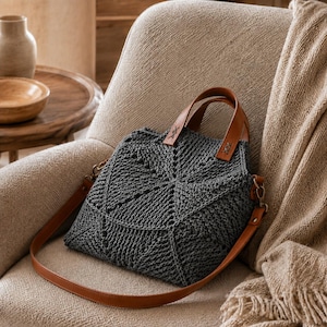 Puede incluir: Bolso tote tejido en crochet gris oscuro con asas de cuero marrón y una correa larga para el hombro. El bolso está sobre un sillón beige con una manta a juego. El bolso tiene un diseño geométrico. Dimensiones: 30 cm x 30 cm.