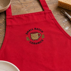 Könnte beinhalten: Eine leuchtend rote Schürze mit einem gestickten Design in der Mitte. Die Stickerei zeigt eine hellbraune Cartoon-Tasse mit einem lächelnden Gesicht, umgeben von dem Text "SMALL BATCH CERAMICS" und kleinen grünen Blumenzweigen mit gelben Akzenten.