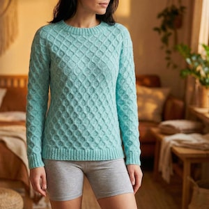 May include: A light blue sweater with a textured diamond pattern. The sweater has a crew neck and long sleeves. The bottom and cuffs have a ribbed design. The person is wearing gray shorts.