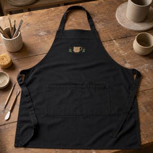 May include: A black apron featuring a central front pocket and a small embroidered graphic on the chest. The graphic depicts a tan ceramic mug with a smiling face, flanked by two green leafy sprigs, with the text Small Batch Ceramics.