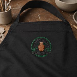 Peut inclure: Un tablier noir avec un logo circulaire brodé en vert et marron. Le logo représente un vase marron central avec le texte POTTERY STUDIO et HANDMADE CERAMICS, entouré de petits motifs de feuilles vertes et de lignes circulaires.