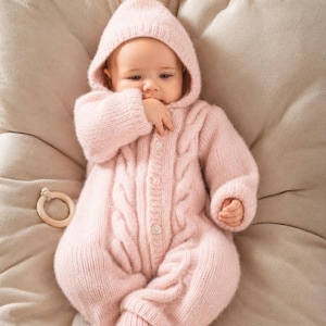 Può includere: Un bambino indossa una tuta in maglia con cappuccio rosa chiaro con un motivo a trecce. La tuta ha una chiusura con bottoni e maniche lunghe. Il bambino è su una superficie beige.