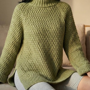 Può includere: Un maglione lavorato a maglia verde chiaro con collo alto e maniche lunghe. Il maglione presenta un motivo a rombi e uno spacco sull'orlo. La modella indossa pantaloncini grigi.
