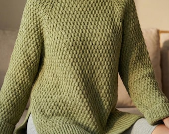 Pode incluir: Um suéter de malha verde claro com gola alta e mangas compridas. O suéter tem um padrão de losangos e uma fenda na bainha. A modelo está usando shorts cinza.
