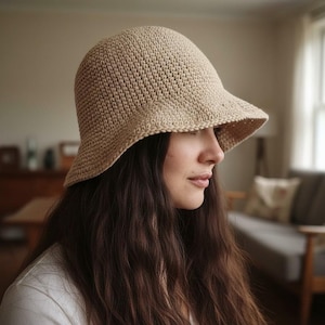May include: A beige crochet bucket hat with a slightly downward-sloping brim. The hat has a textured appearance and a rounded crown. The hat is made of a light-colored yarn and is suitable for sun protection.